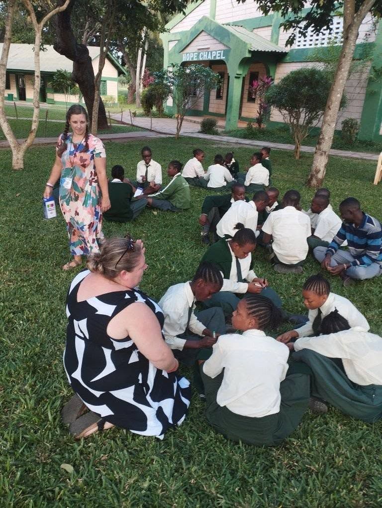 Children and two adults sit on grass in a garden, with a woman standing nearby and a small building in back.