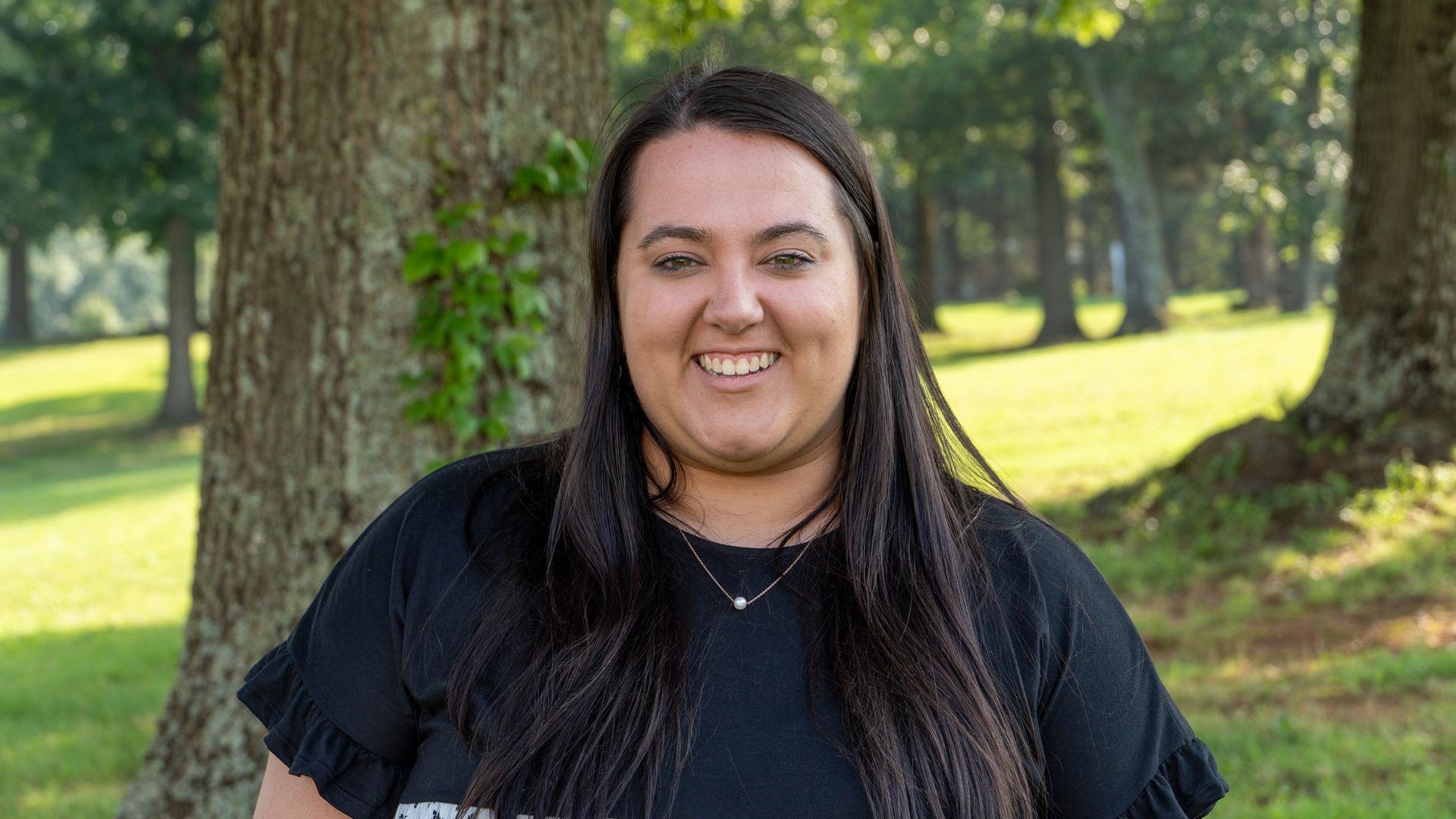 Smiling person in a black shirt standing outdoors near a tree in a grassy park