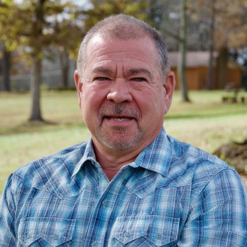 Man in a blue plaid shirt standing outdoors with trees and a grassy yard in the background