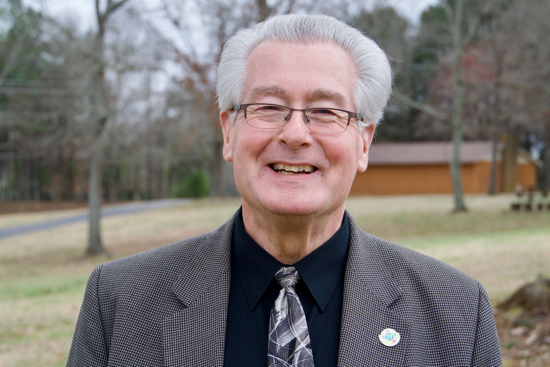 Smiling man in a patterned jacket standing outdoors near a road and trees