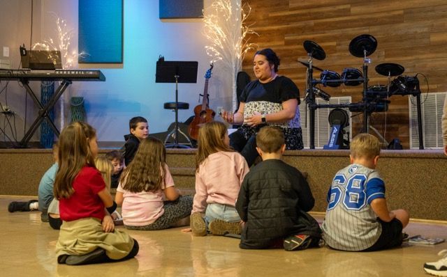 Children sit on a stage floor listening to a speaker beside a microphone and drum kit.