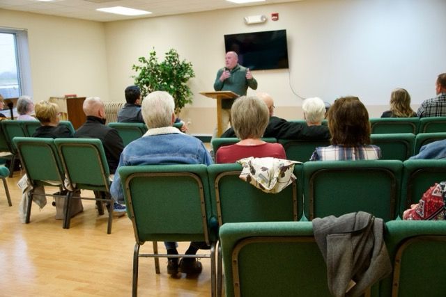 Small meeting room with seated audience listening to a speaker beside a wall-mounted TV.