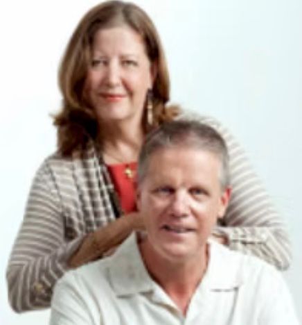 Two smiling adults posed against a white background, the woman standing behind the seated man.