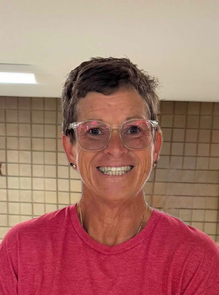 Smiling person in pink shirt and clear glasses standing indoors against tiled wall