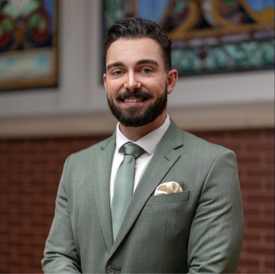 Man in a sage green suit smiling indoors, with framed art and brick wall in the background