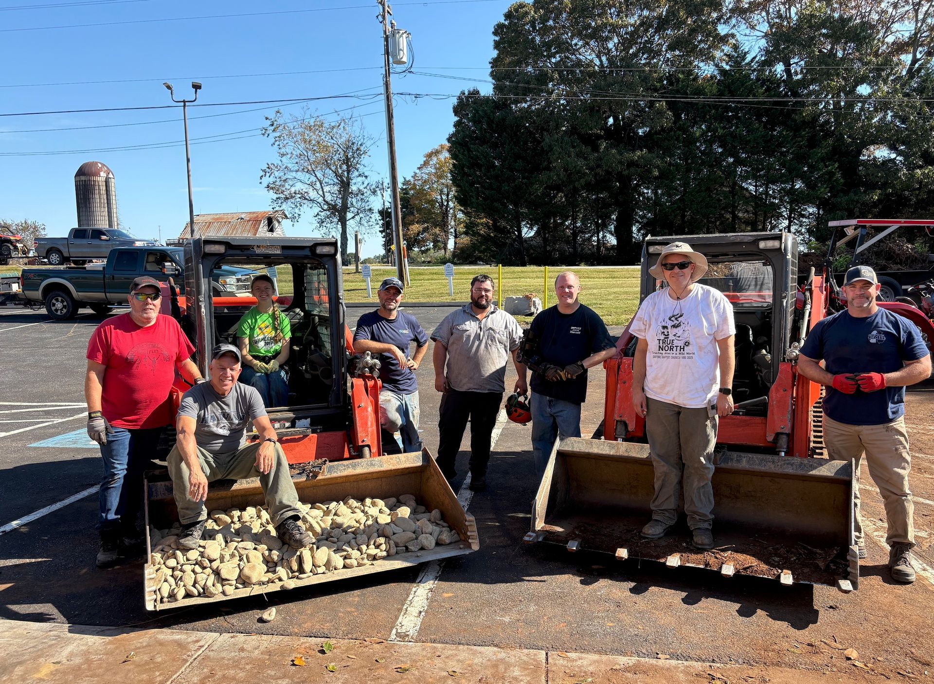 Group standing by wooden barriers and tools in a sunny parking lot, with tractors and trees in the background.