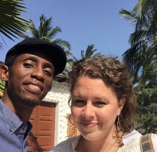 Two people smiling for a selfie in front of a house with palm trees.