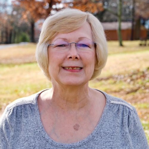 Smiling person with glasses standing outdoors in a park, wearing a gray top.