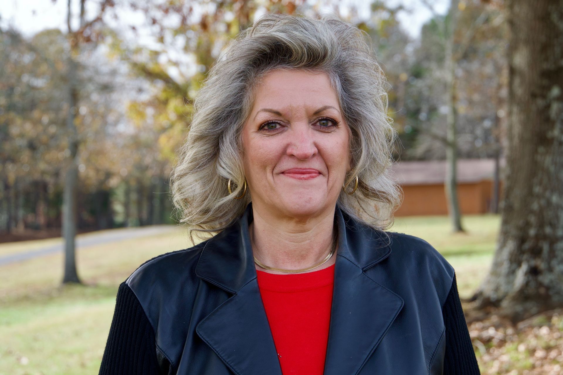Woman in a black jacket and red top stands outdoors in a park, with trees and grass in the background.