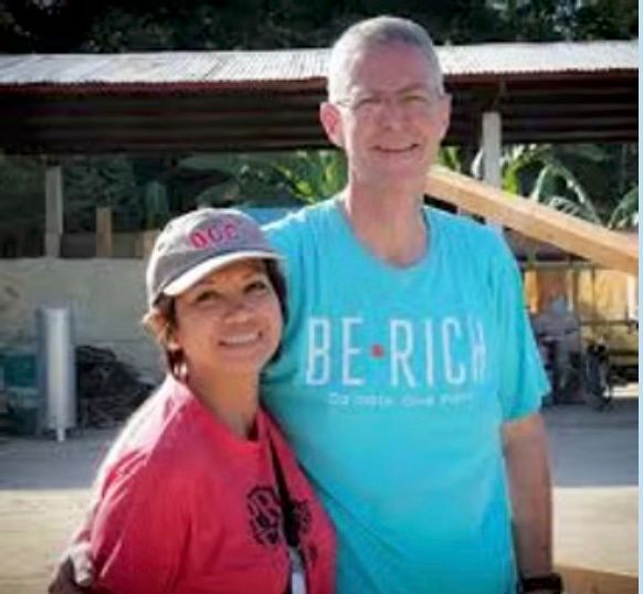Two people smiling outdoors; one wears a turquoise BE-RICH T-shirt and the other a pink shirt and cap.
