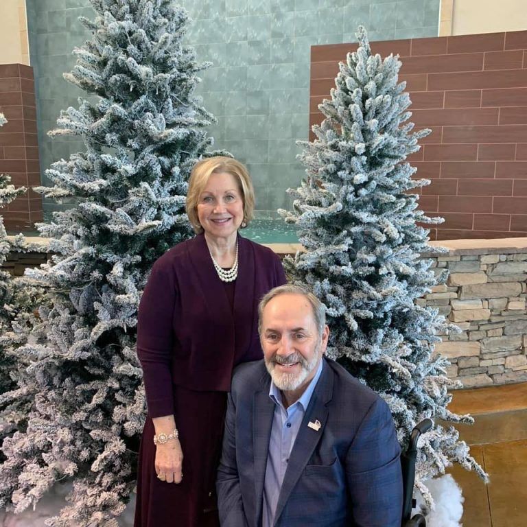 Two people posing indoors in front of snow-dusted Christmas trees and a stone wall.