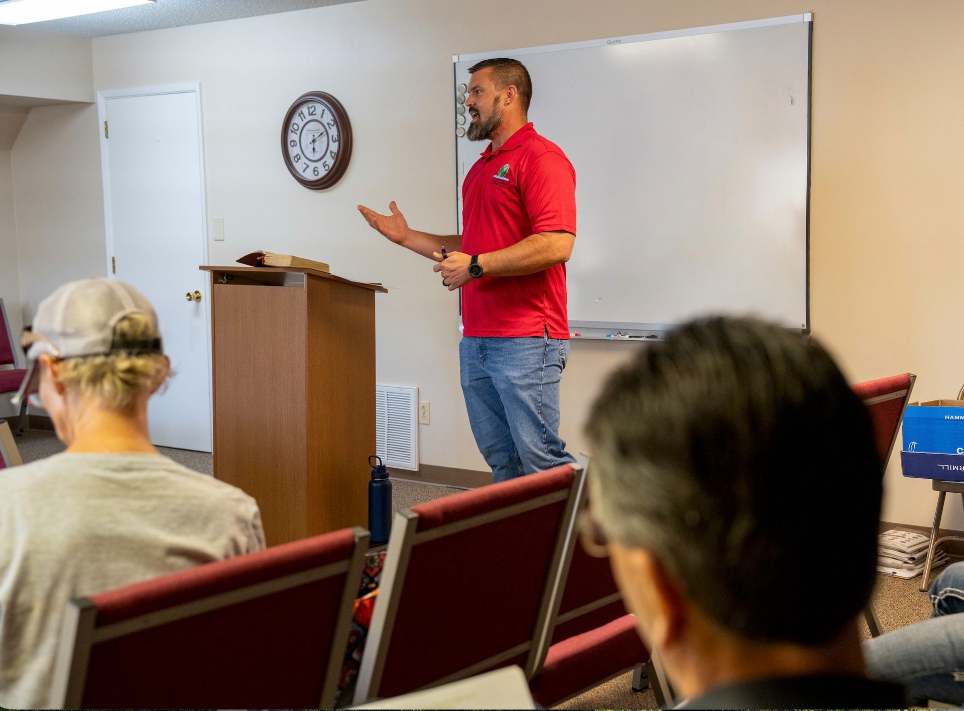 Man speaking beside a podium in a small classroom, with seated audience and whiteboard behind him.