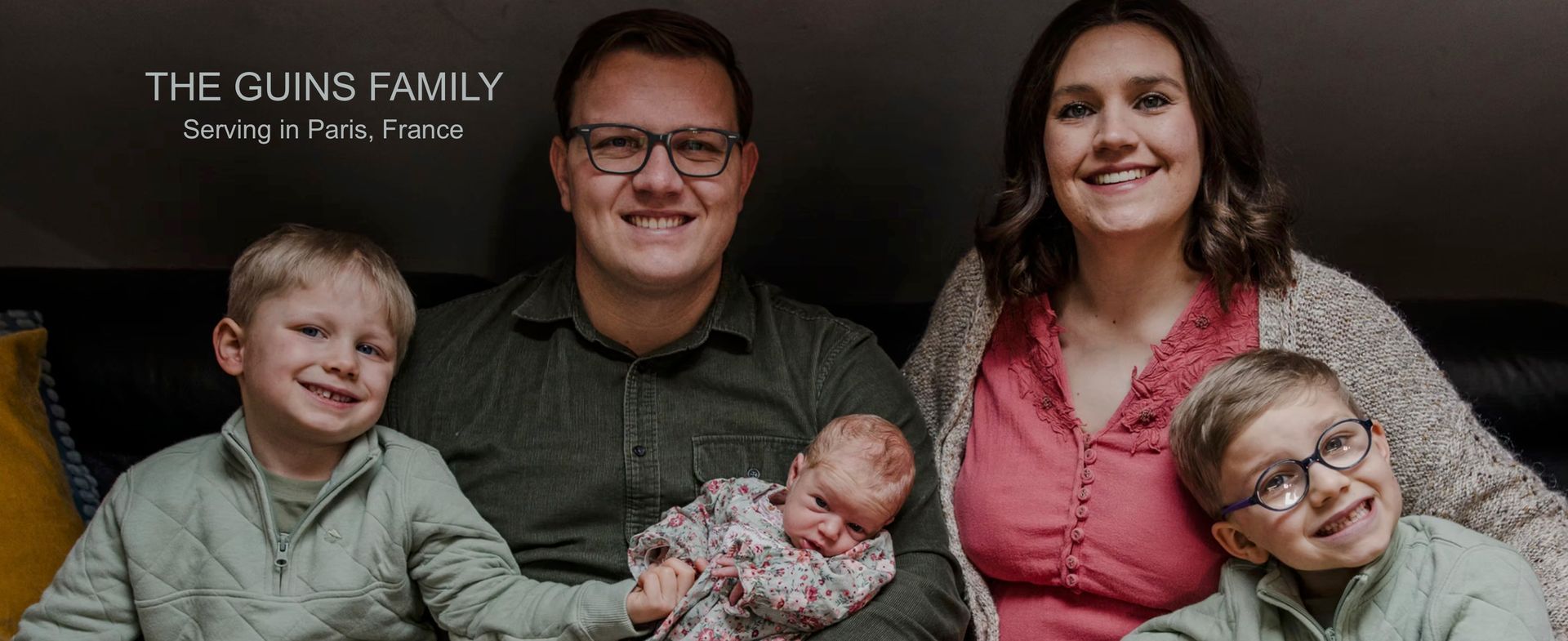 Family portrait of five smiling people on a couch, with a baby wrapped in a blanket at center.