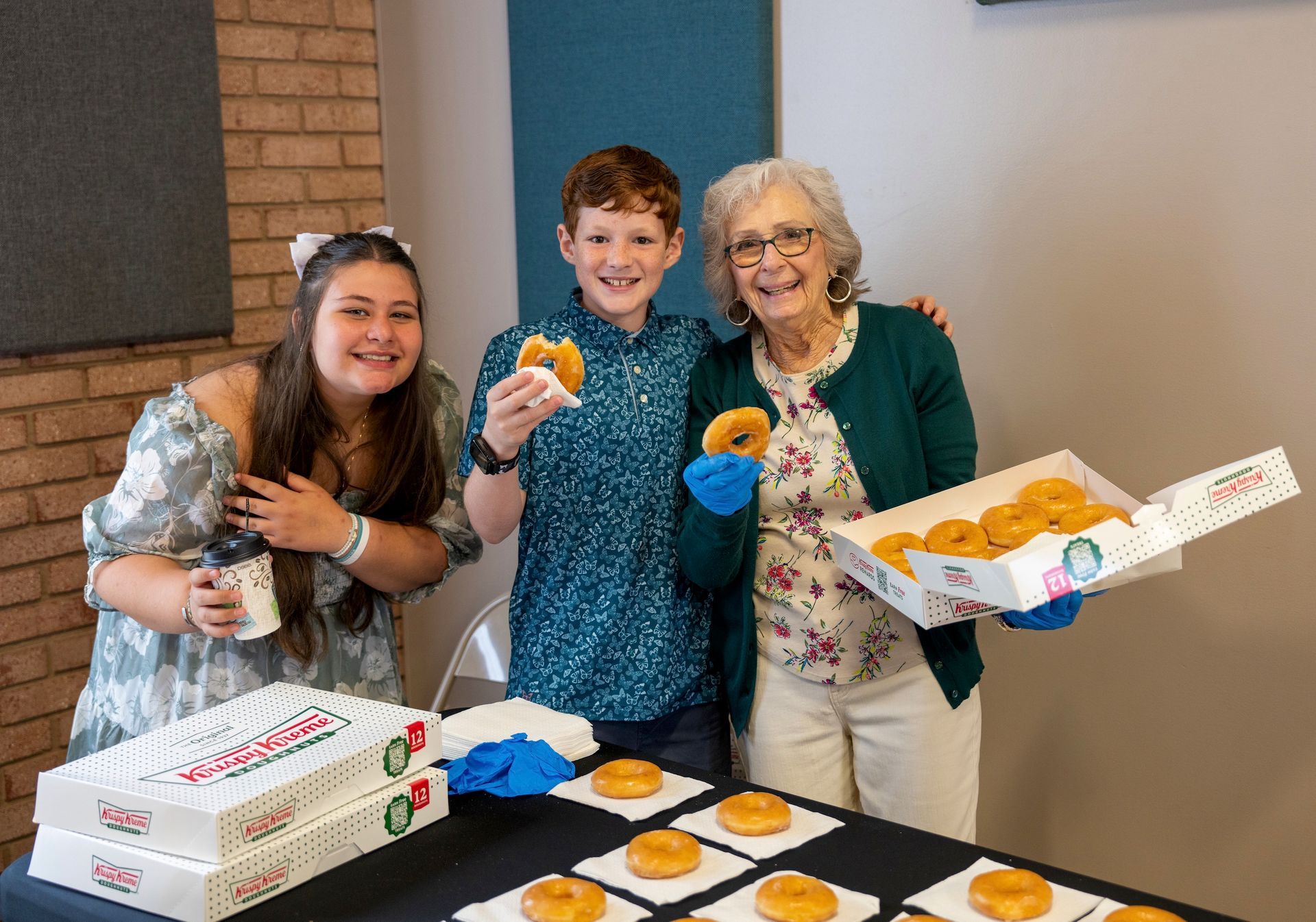 Three people holding cupcakes beside an open box and tray of donuts at a table