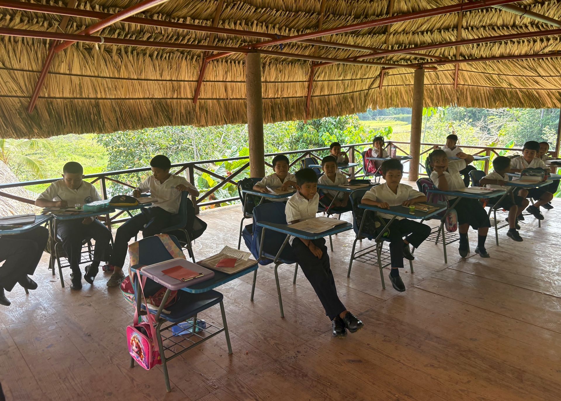 Students seated at desks in an open-air thatched classroom, facing forward during a lesson.