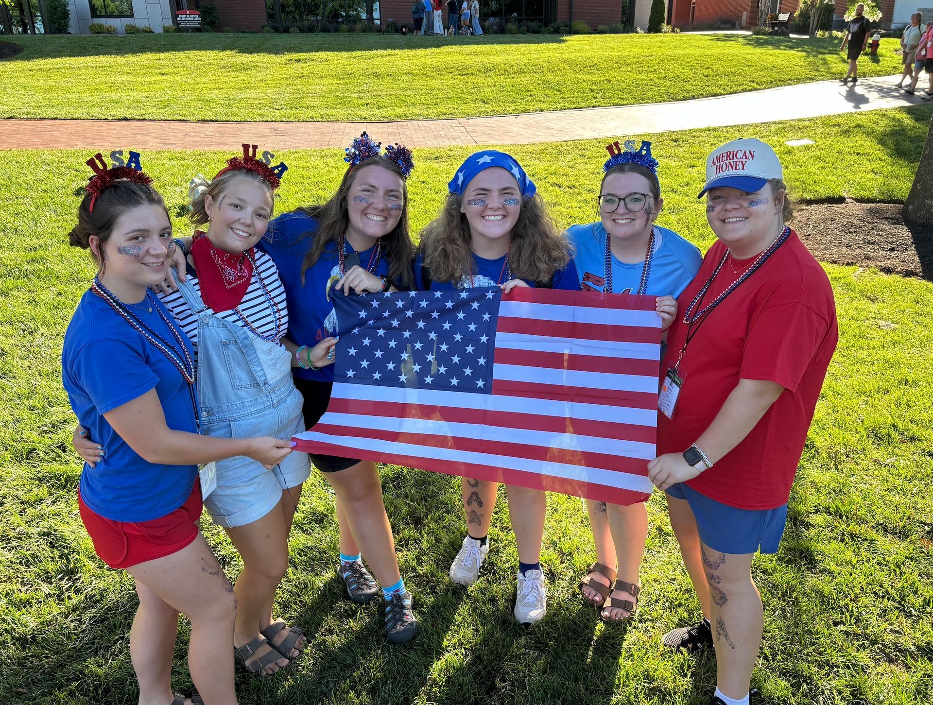 Six people holding an American flag on a grassy lawn in bright sunlight