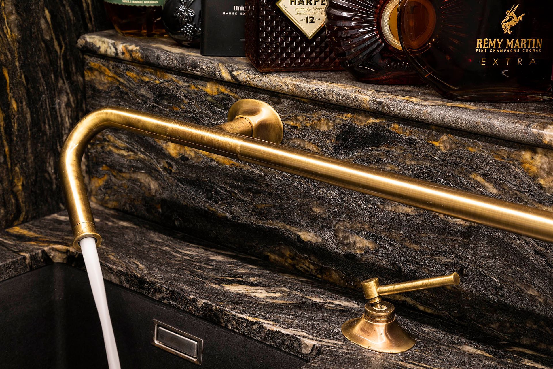 Brass outdoor shower fixture with a curved gold pipe and white shower handle against dark stone tiles — Shona O'Brien Interior Design in Bowral, NSW