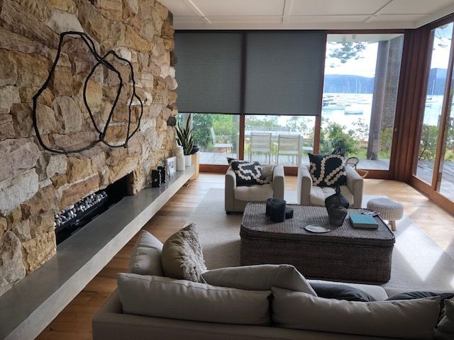 Modern living room with stone fireplace, gray sofas, woven coffee table, and large windows overlooking water — Shona O'Brien Interior Design in Bowral, NSW