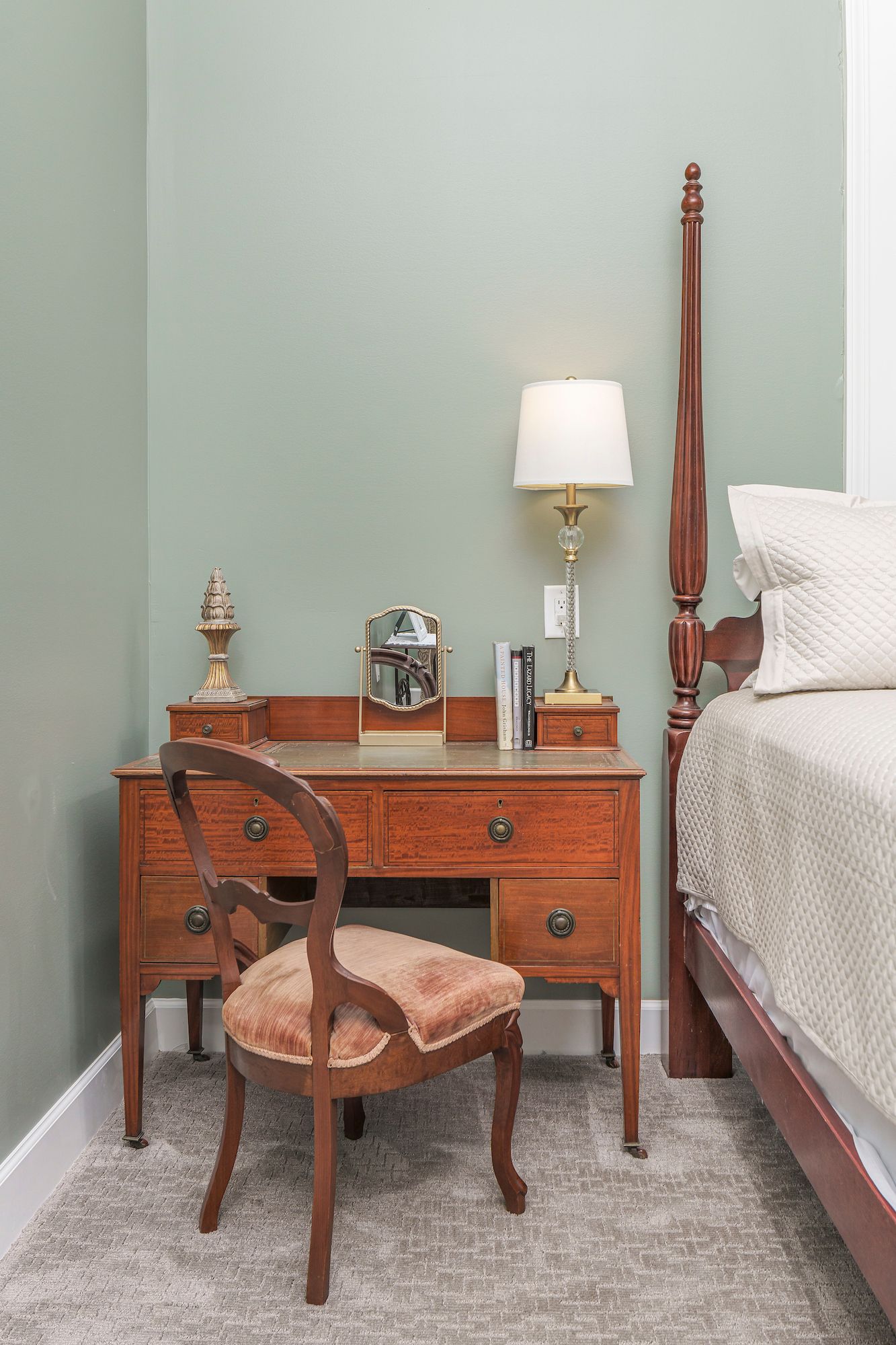 Bedroom corner with wooden desk, chair, and bed. Lamp and mirror on desk against a green wall.