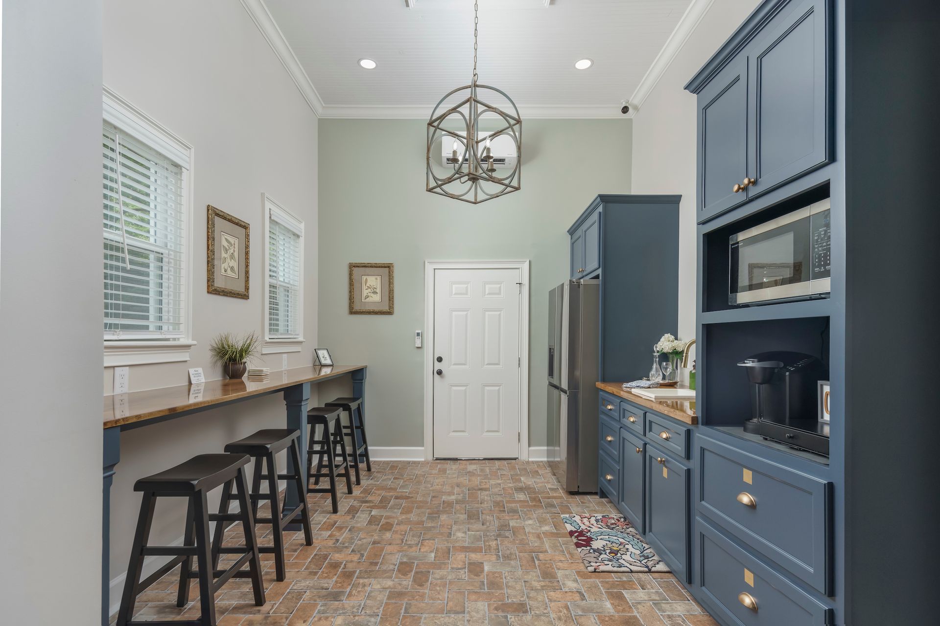 A narrow kitchen with blue cabinets, a breakfast bar with stools, and a brick floor.
