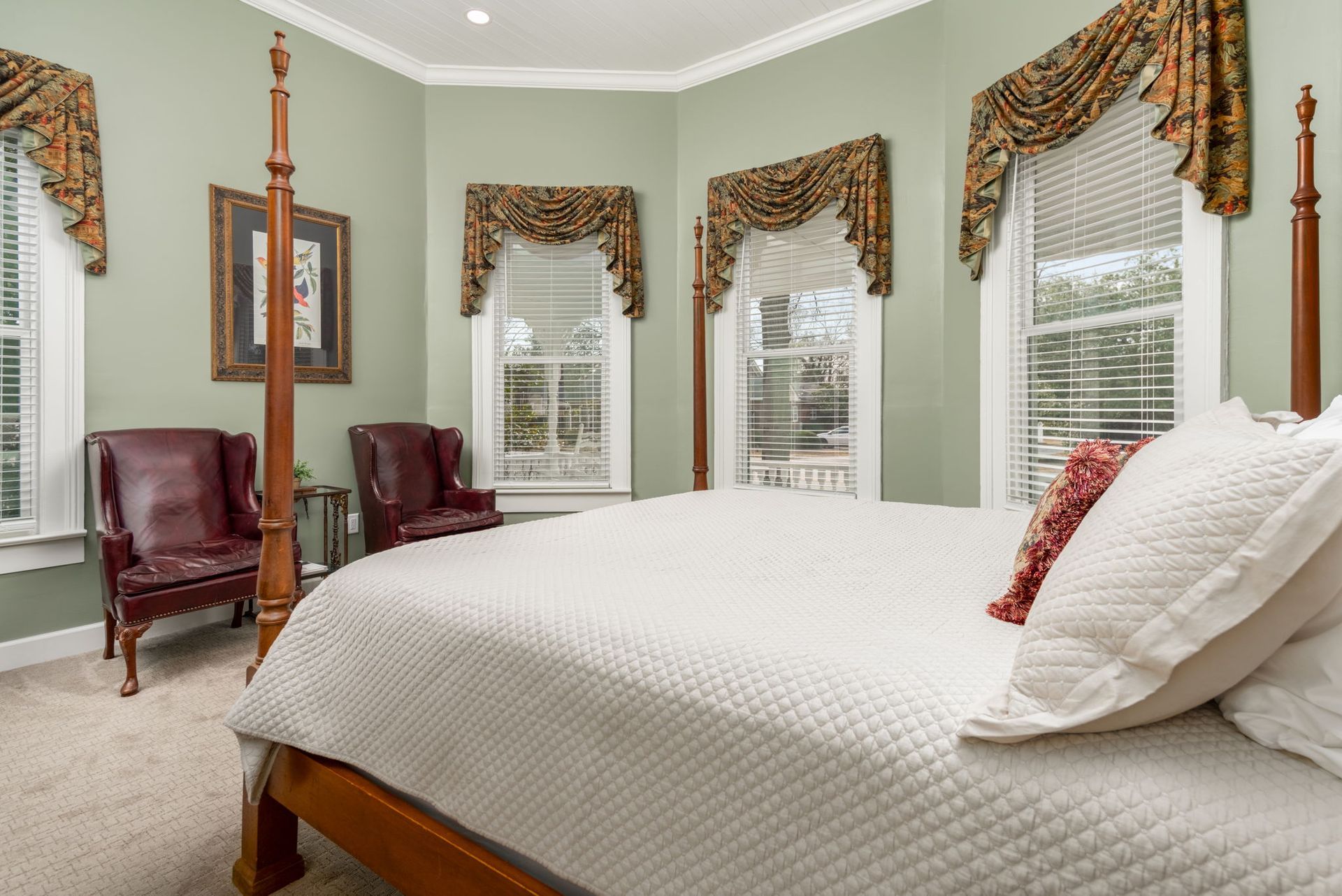 Bedroom with four-poster bed, two red chairs, three windows with curtains, and green walls.