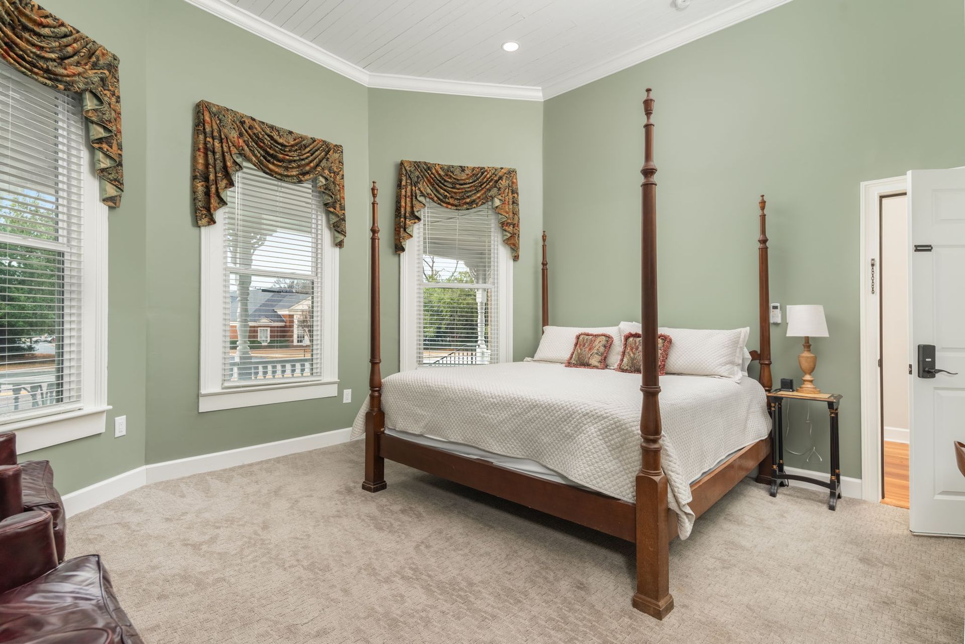Bedroom with four-poster bed, sage green walls, three windows with ornate drapes, and a neutral carpet.