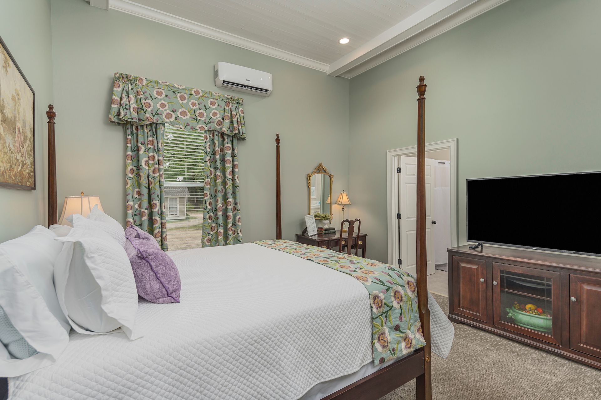 Bedroom with four-poster bed, floral drapes, TV, vanity, light green walls, and white bedding.