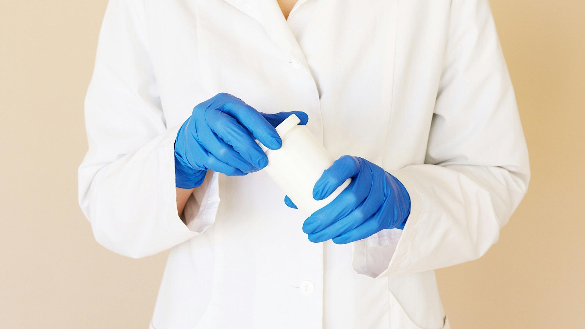 A woman in a lab coat and blue gloves is holding a white bottle.