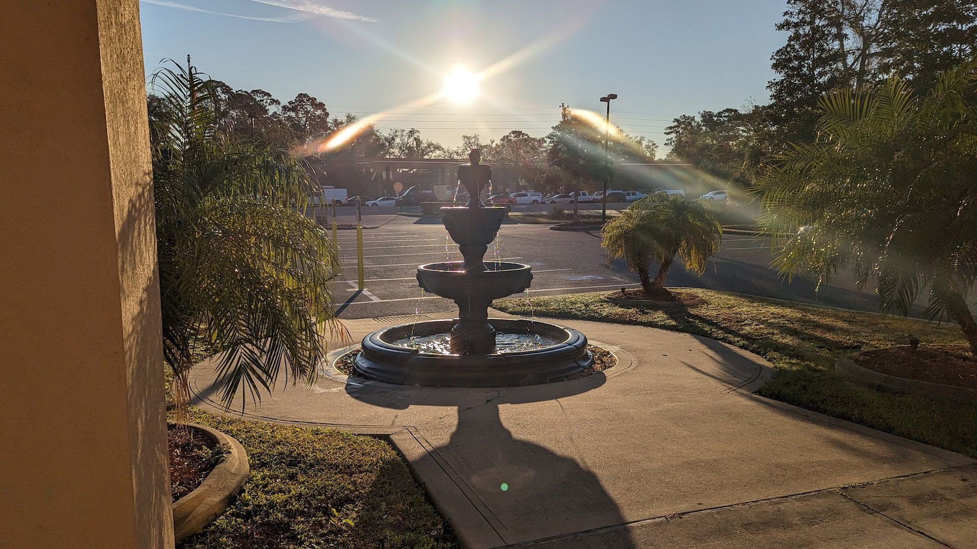 Fountain in sunlight casts shadow on walkway, trees surround, parking lot in background.