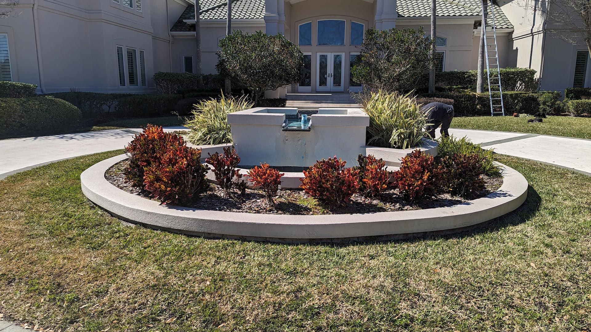 Circular flower bed with shrubs in front of a house, person working on the water fountain in the background.
