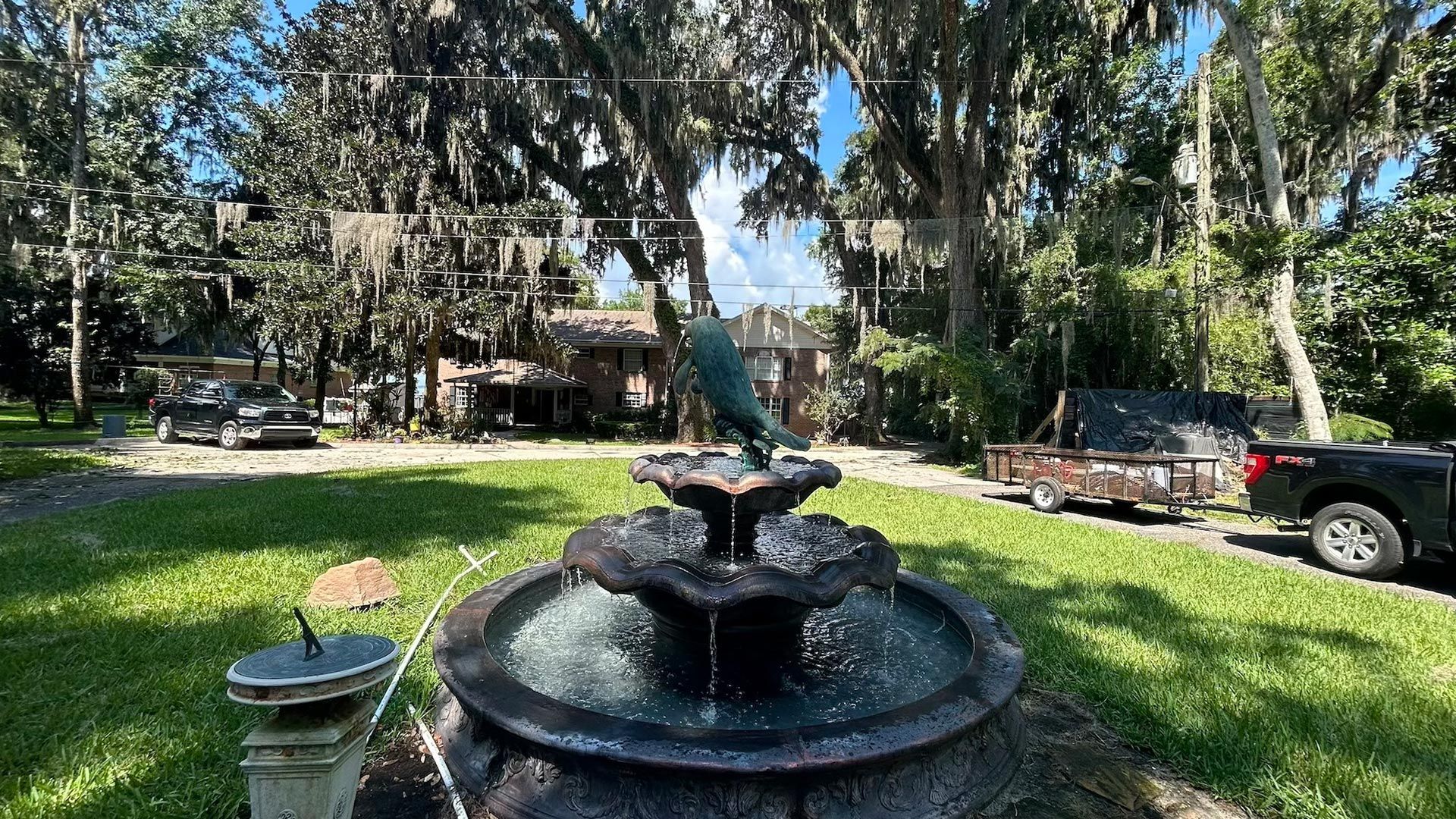 Fountain with bird statue, house in background, green lawn, trees, blue sky.