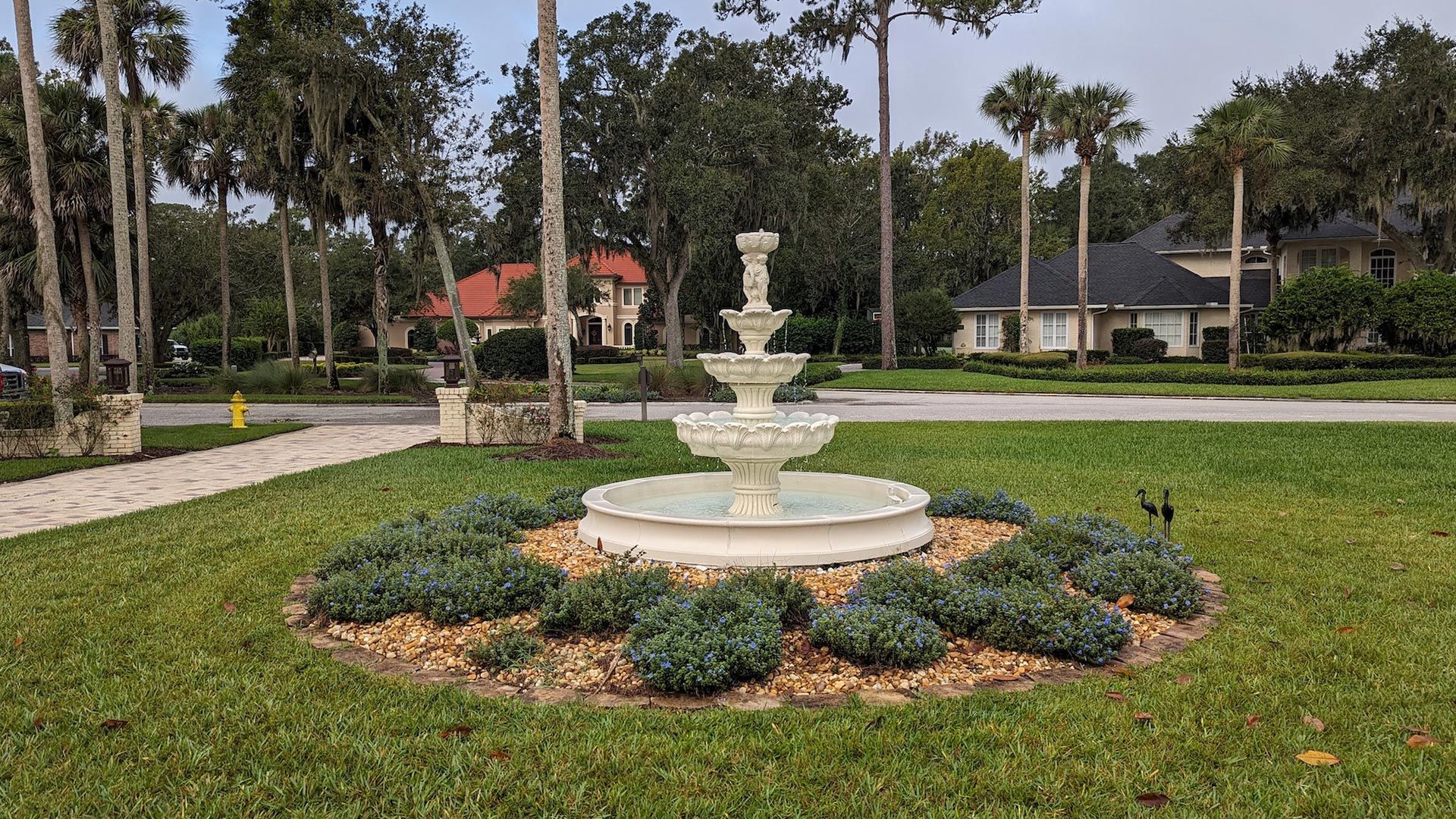 White tiered fountain surrounded by green shrubs, in a grassy area with trees and houses.