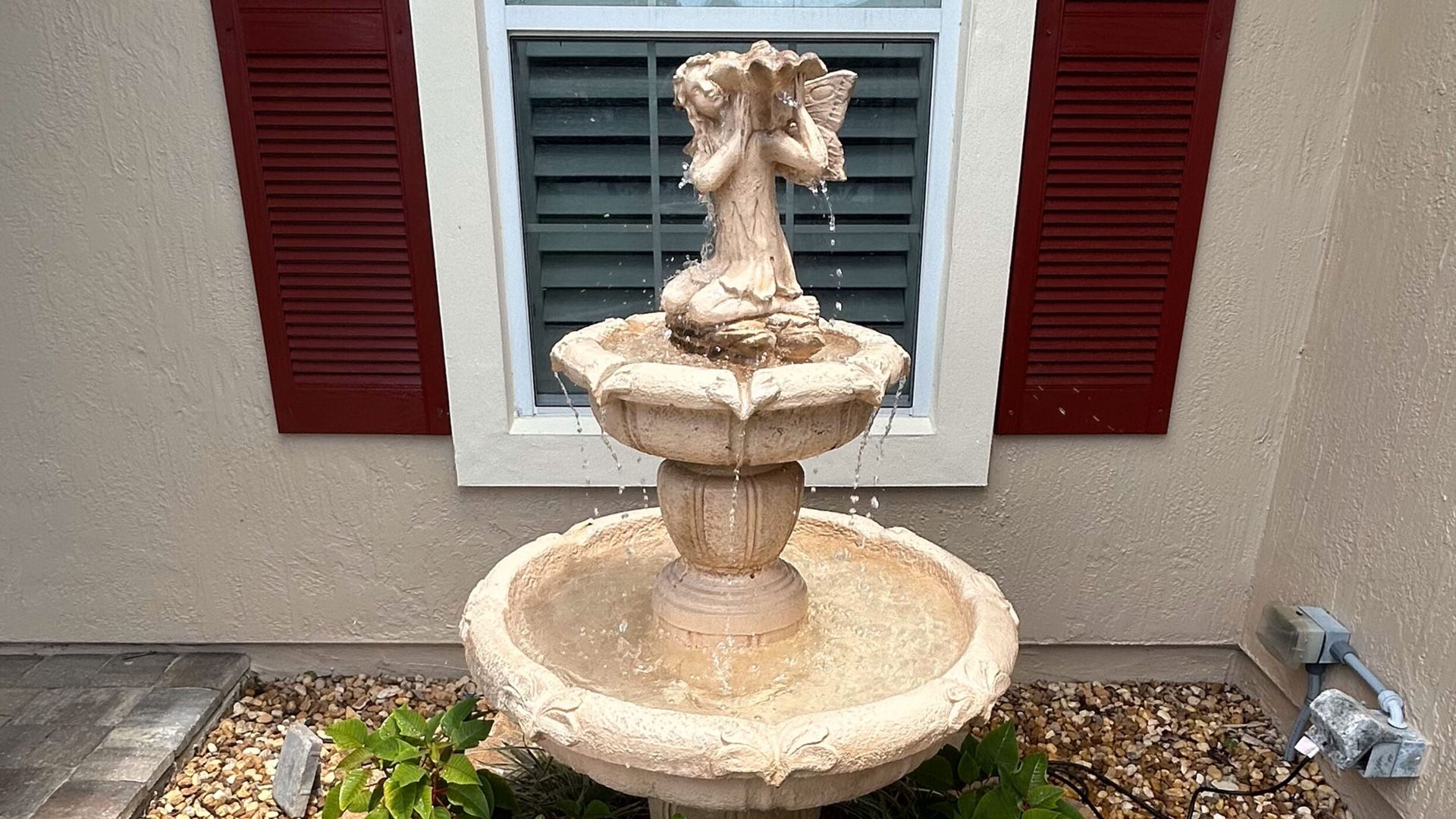 Water fountain with two-tiered basins and angel statue, beige, in front of a window with red shutters.
