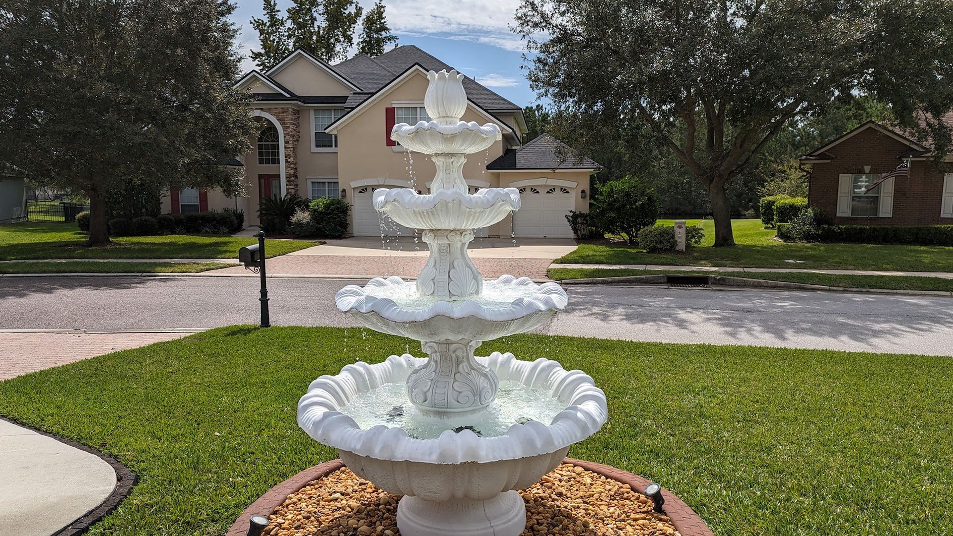 White tiered fountain in front yard with house in the background. Green grass, blue sky.