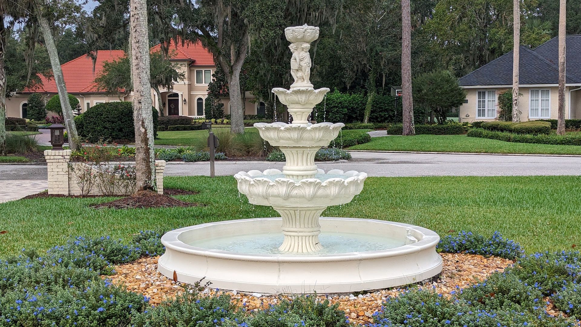 Stone fountain with tiered bowls in a grassy yard, with houses and trees in the background.