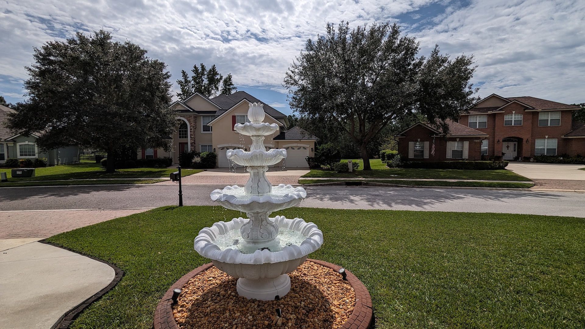 White tiered fountain in a suburban front yard, with houses and trees in the background under a cloudy sky.