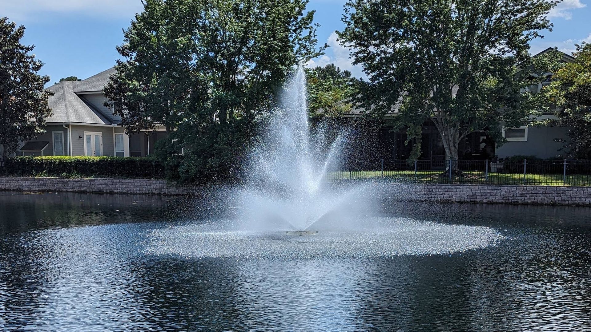 Fountain spraying water in a pond with houses and trees in the background under a blue sky.