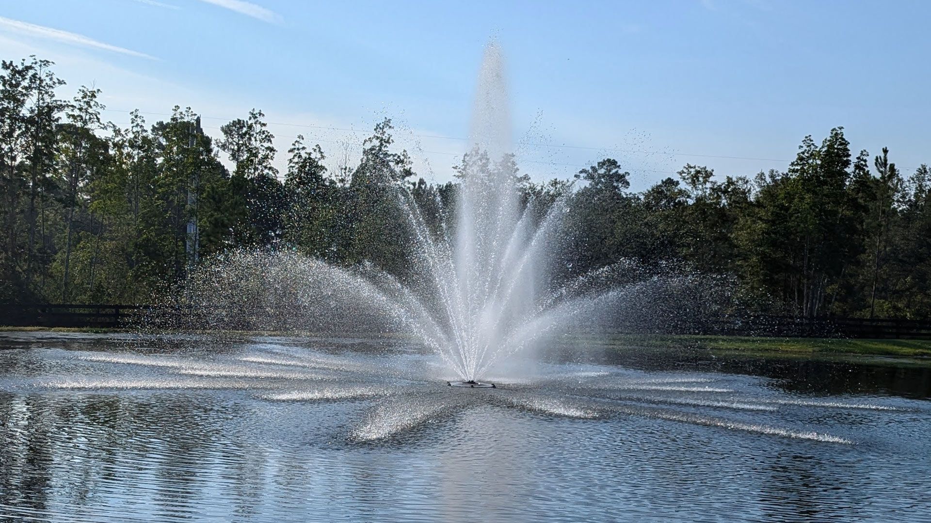 Fountain spraying water high into the air in a pond, with trees and sky in the background.