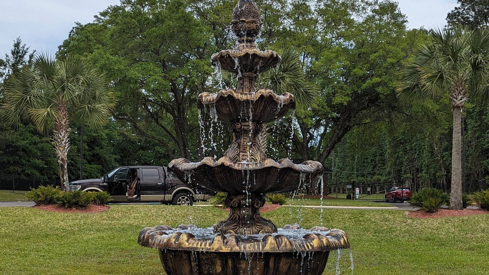 Ornate tiered fountain on green lawn with trees and parked vehicle.