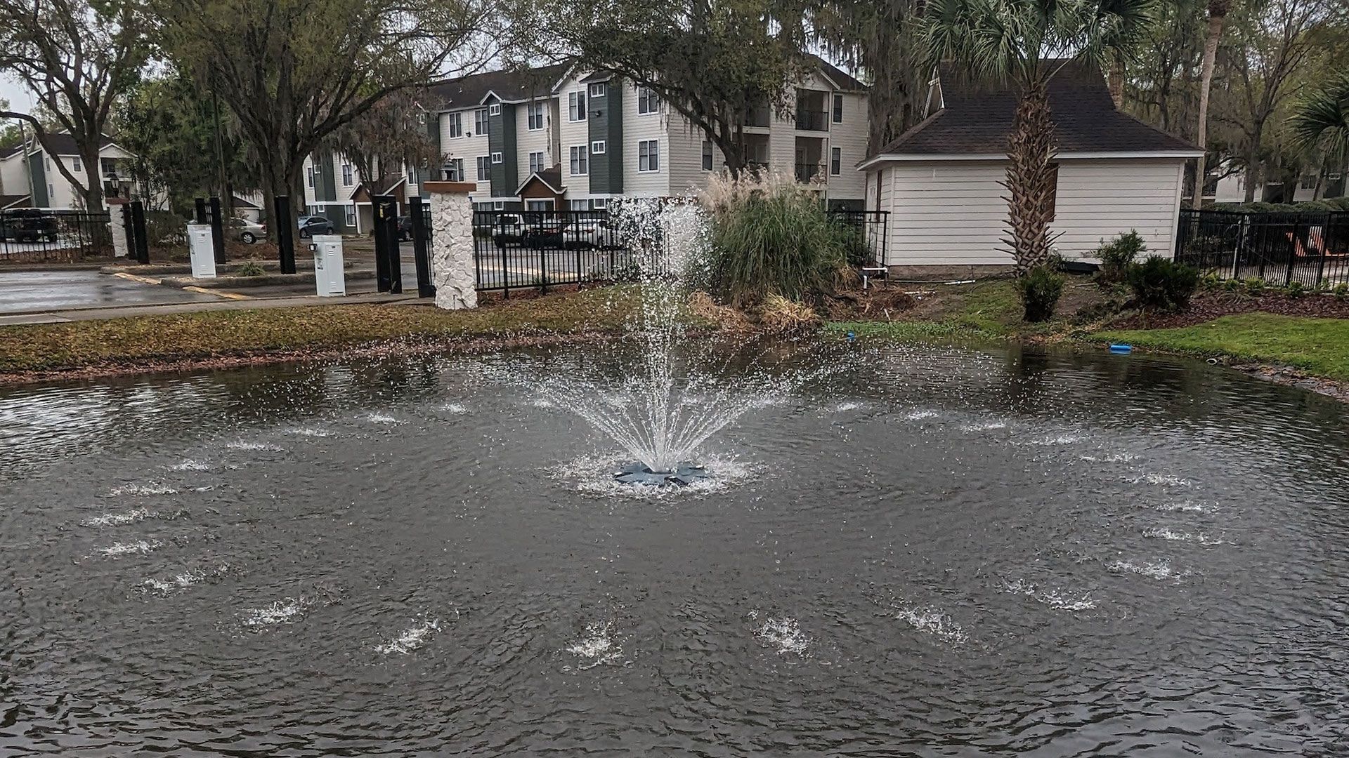 Pond with water fountain, spray jets, and apartment buildings in the background. Two people are standing near a pillar.