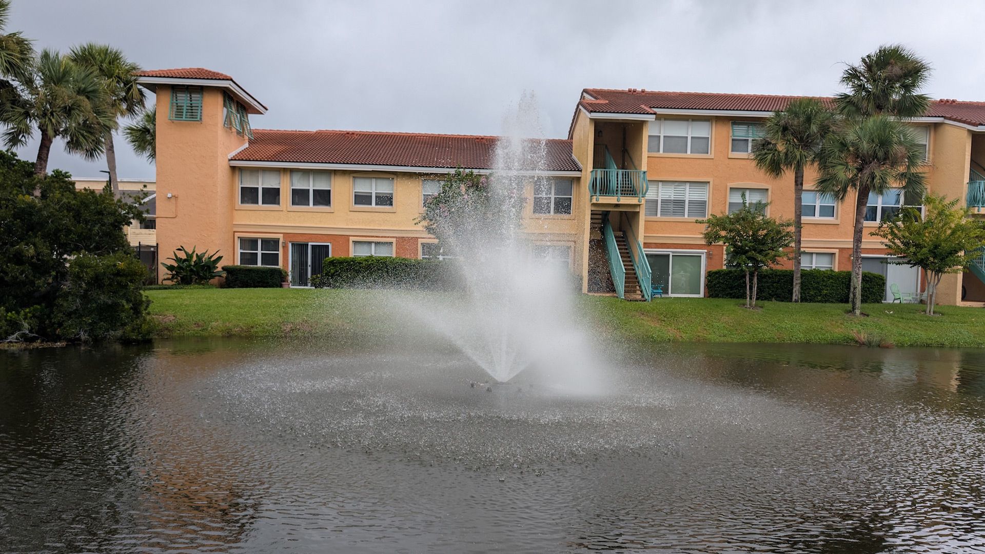 Fountain sprays water in front of an orange apartment building on a cloudy day. Green grass and palm trees surround.