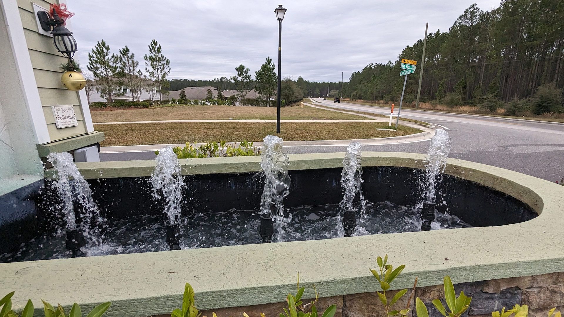 Fountain with water spouts, sign, road, and trees in a residential area on an overcast day.