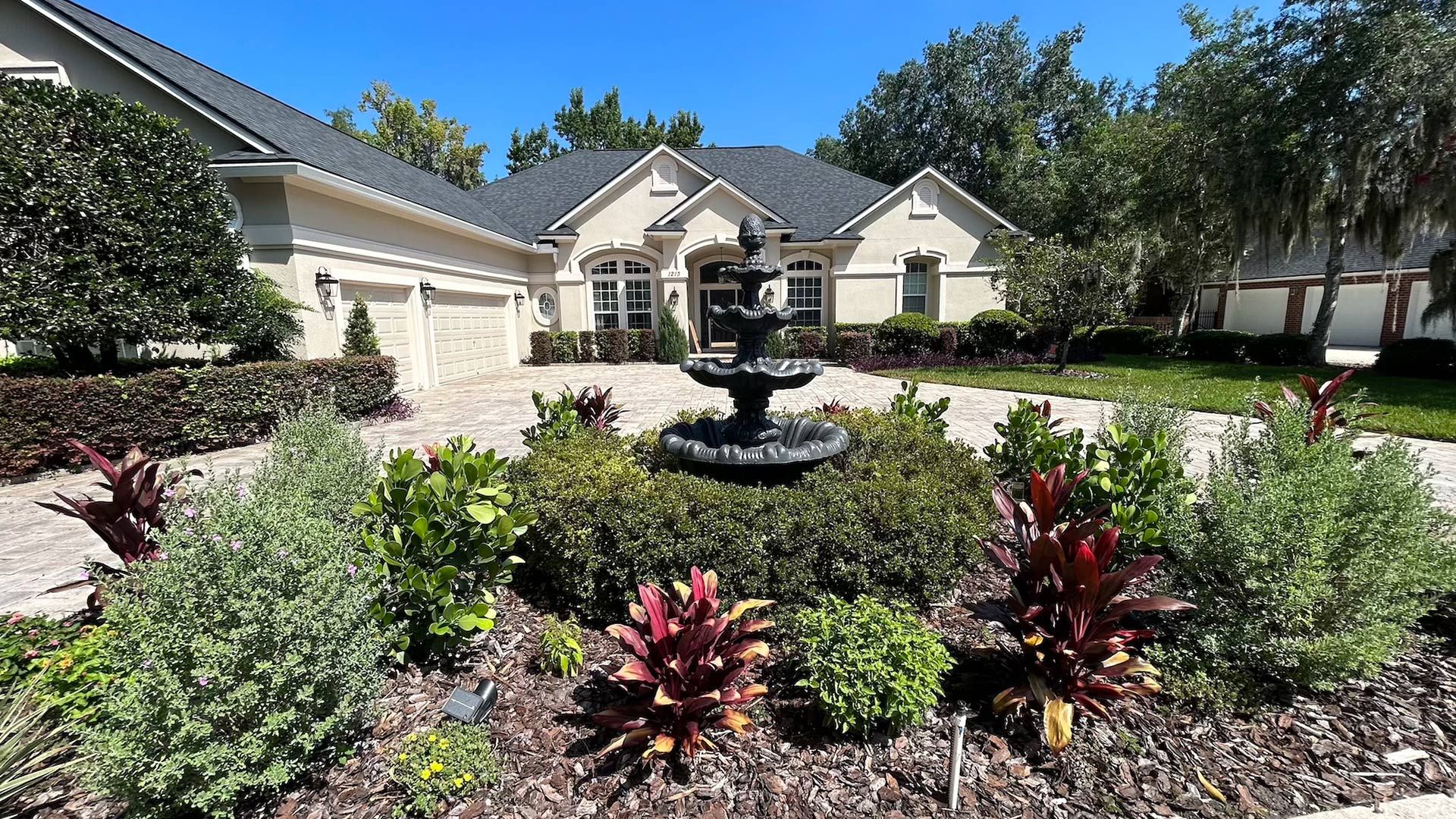 Beige house with a dark roof and a fountain in a landscaped front yard.