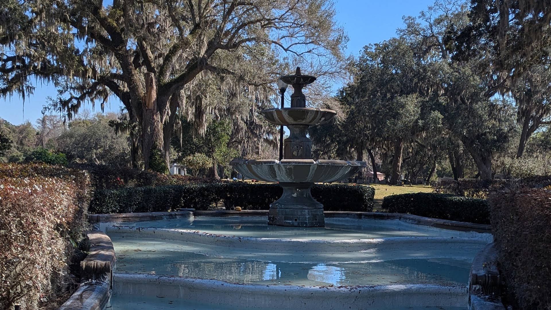 Fountain in a garden setting, surrounded by trees and shrubbery, on a sunny day.