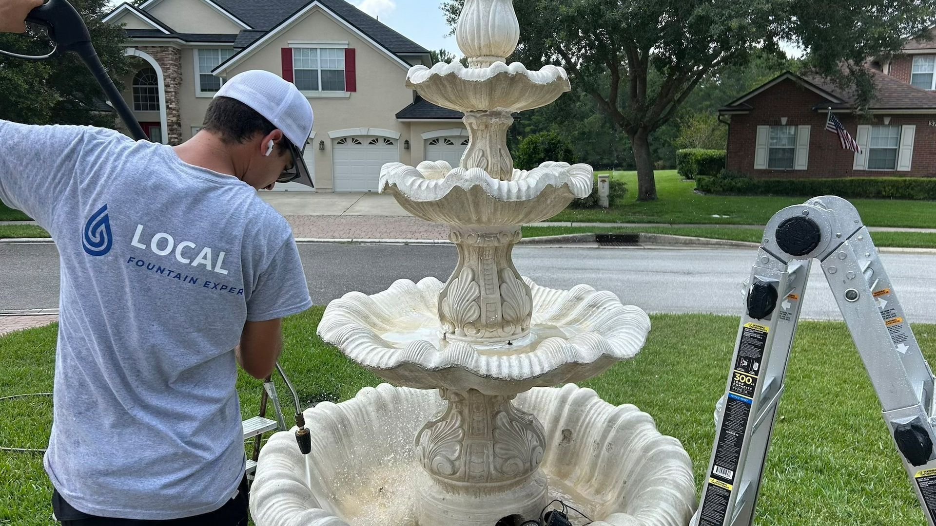 A man in gray shirt power washes a three-tiered fountain on a front lawn with a ladder nearby.