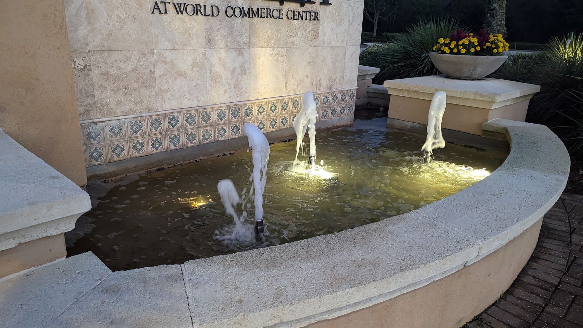 Fountain with water spouts, lit up at night, in front of a building with a flower pot on the side.