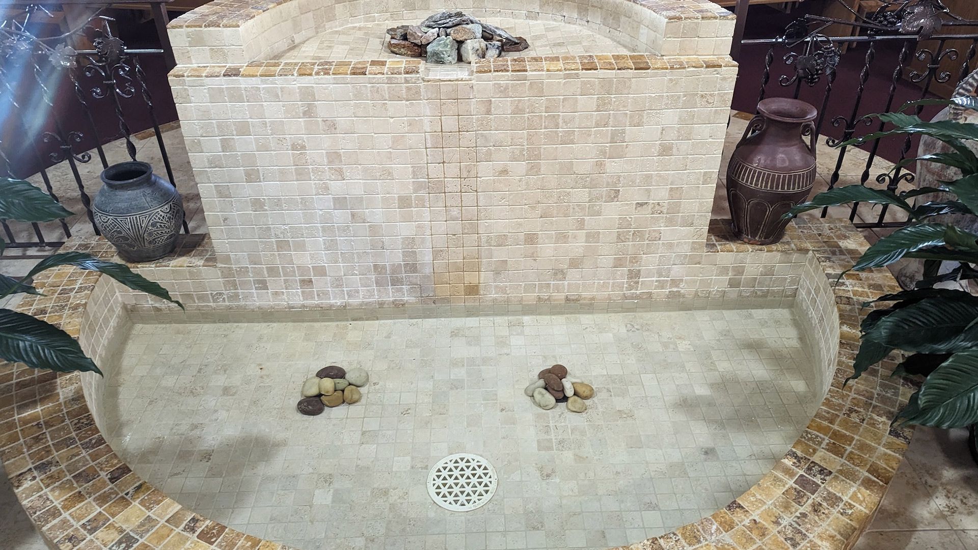 Indoor stone fountain with two levels, jars, and plants.