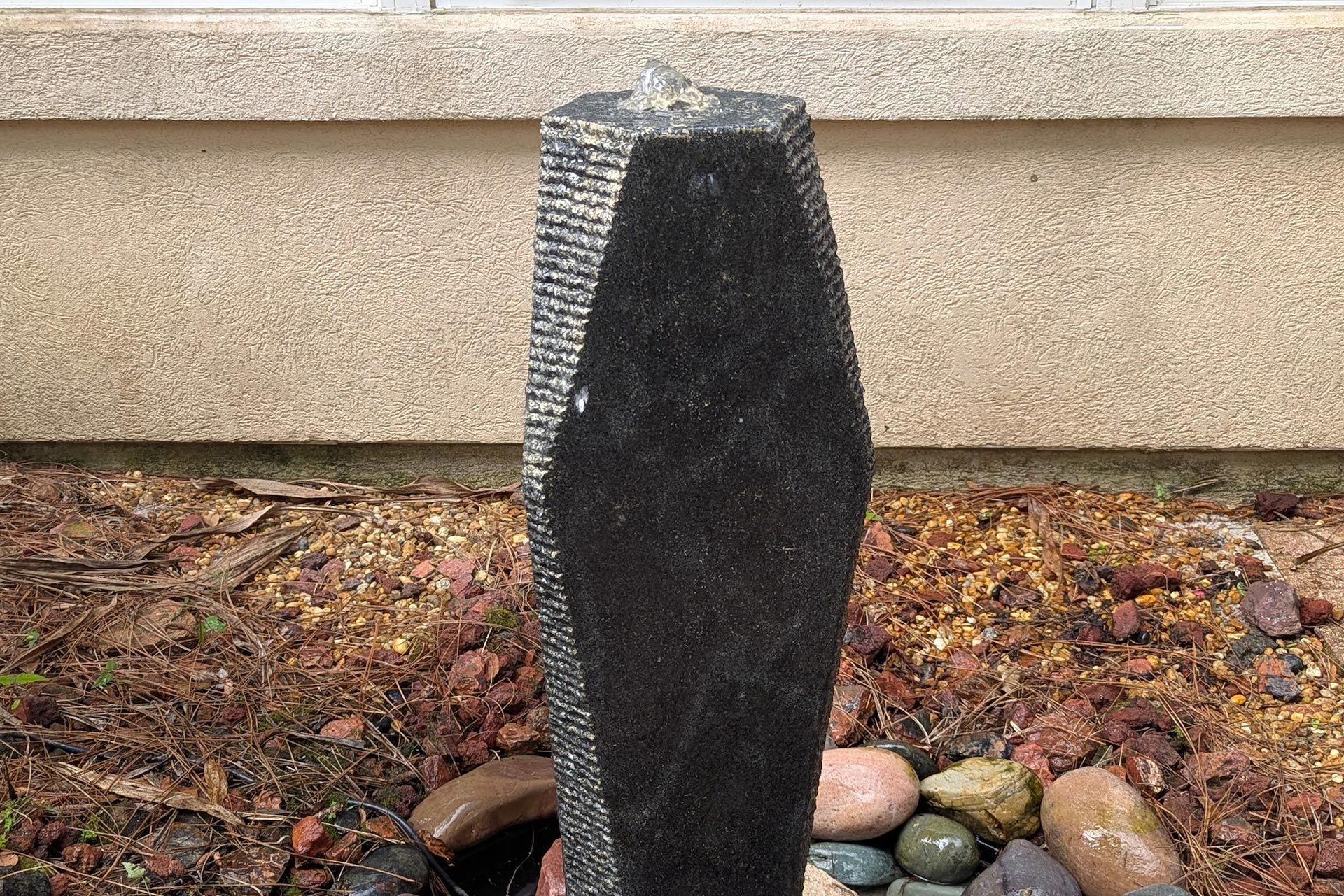 Black stone water fountain with water trickling from top, set in a garden with rocks and soil.