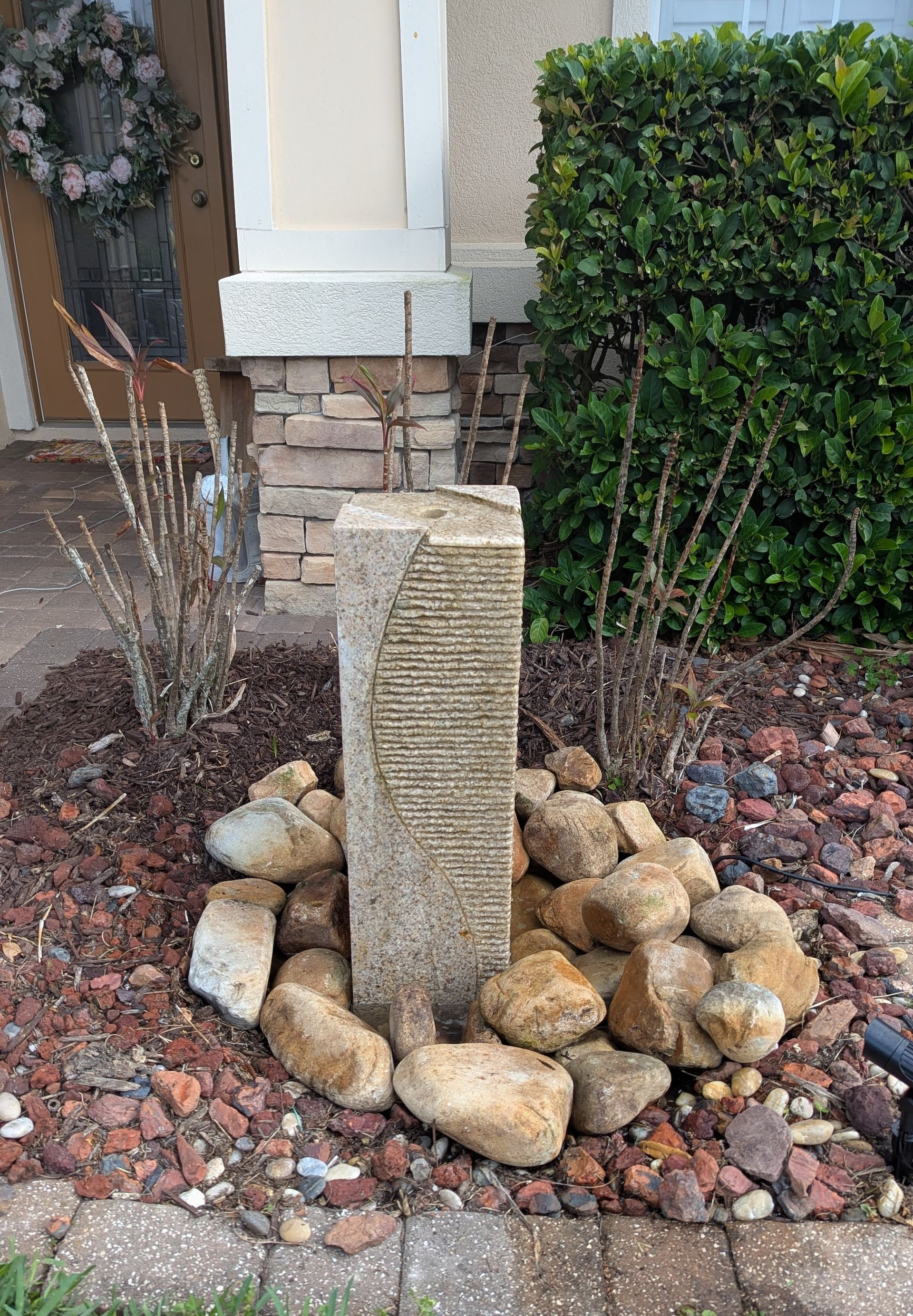 Stone water fountain surrounded by rocks and brown mulch in front of a house.