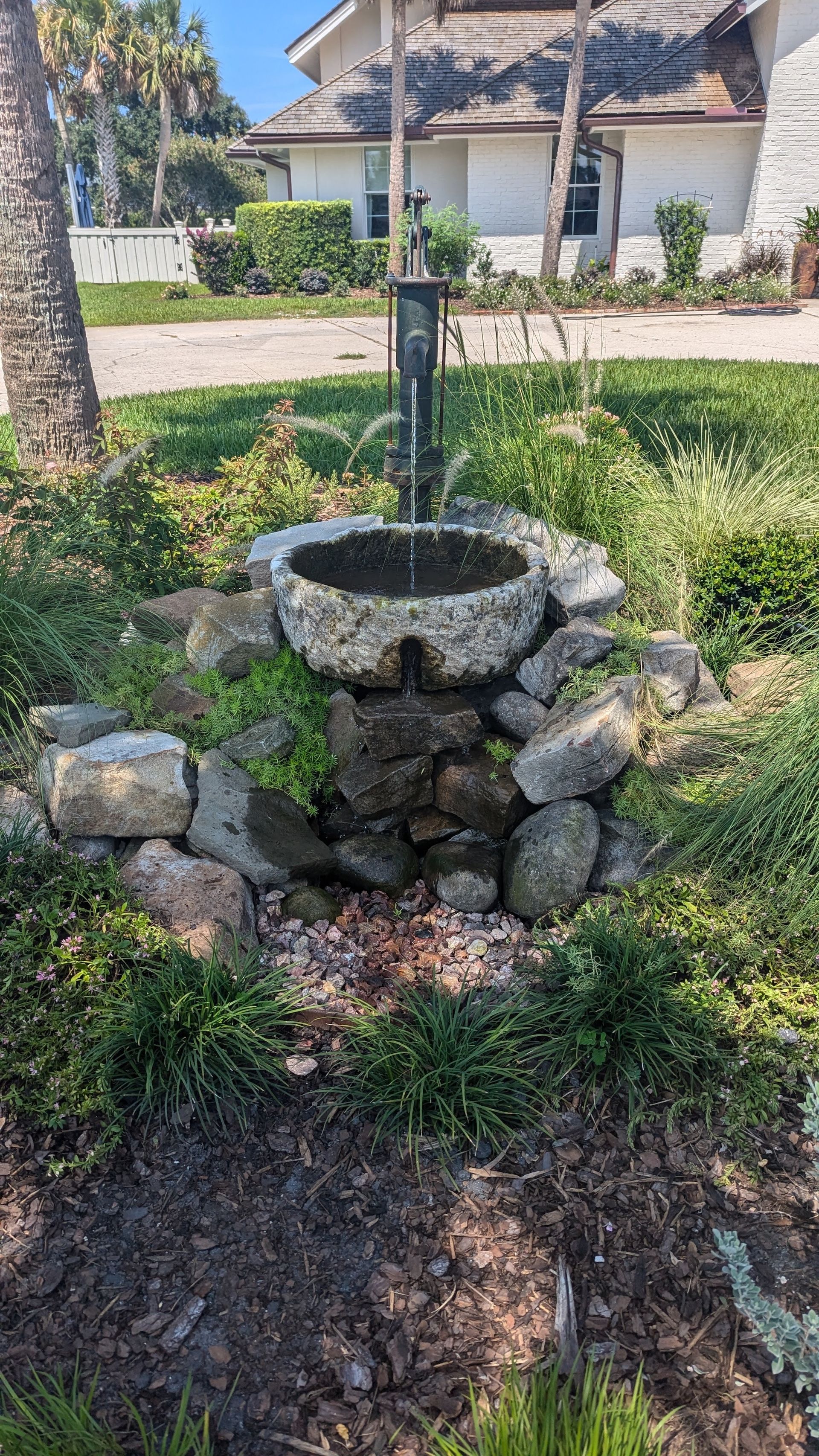 Water fountain in a garden with rocks and green plants, house in the background.